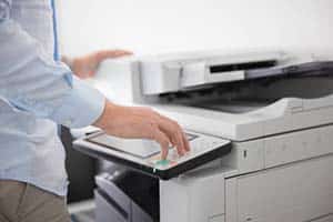 Person operating a photocopier, pressing a button on the control panel with the machine's lid open, as if preparing for a small format printing task.