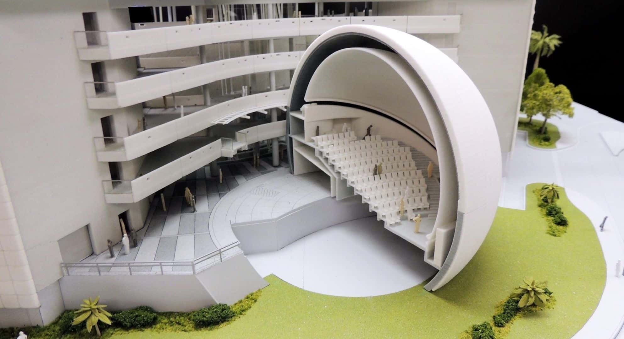         A scale model of a modern amphitheater with tiered seating, partially enclosed by a circular dome, adjacent to a multi-story building, surrounded by green landscaping and pathways. The intricate details are meticulously crafted using 3D printing technology.