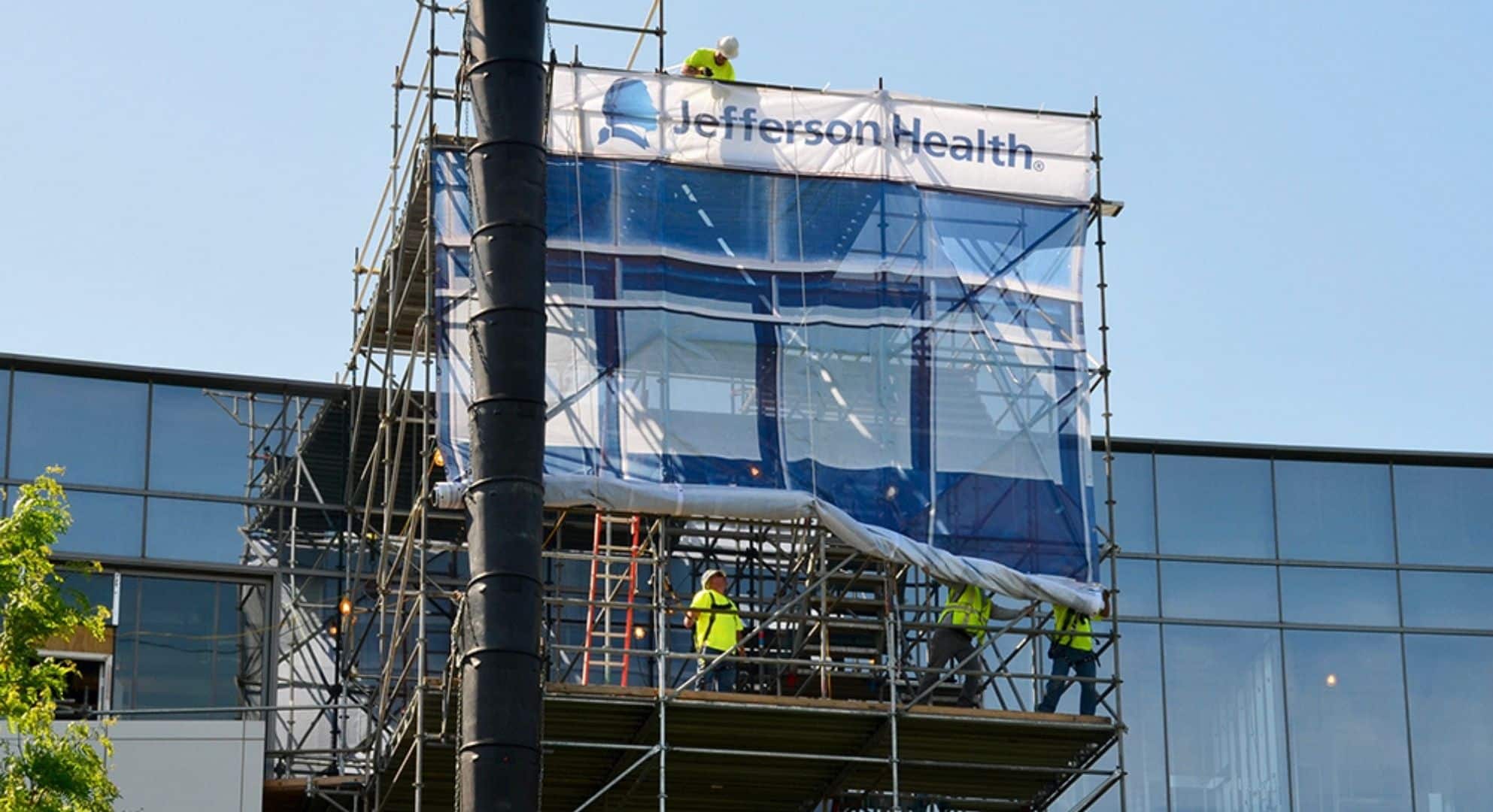 Construction workers are on scaffolding around a building covered with a plastic sheet displaying the 