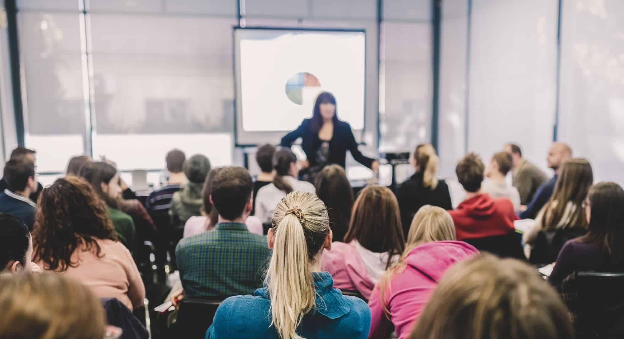 A woman is presenting to a classroom of people, with a projector screen displaying a pie chart in the background. As she discusses market trends, she highlights innovations like large format printing that are transforming visual communication.