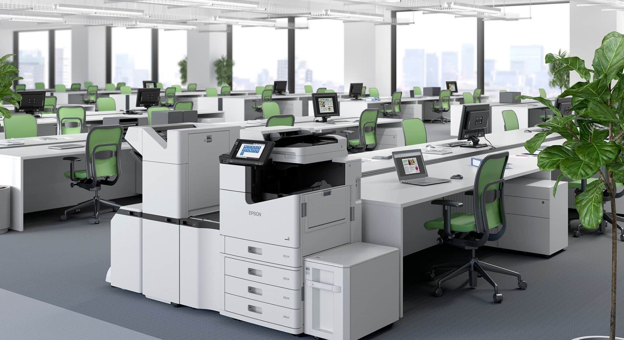 A modern office space with green chairs, white desks, computer monitors, and a large printer/copier in the center caters to all your web-to-print needs. Indoor plants are placed in the background, adding a touch of freshness to the room.