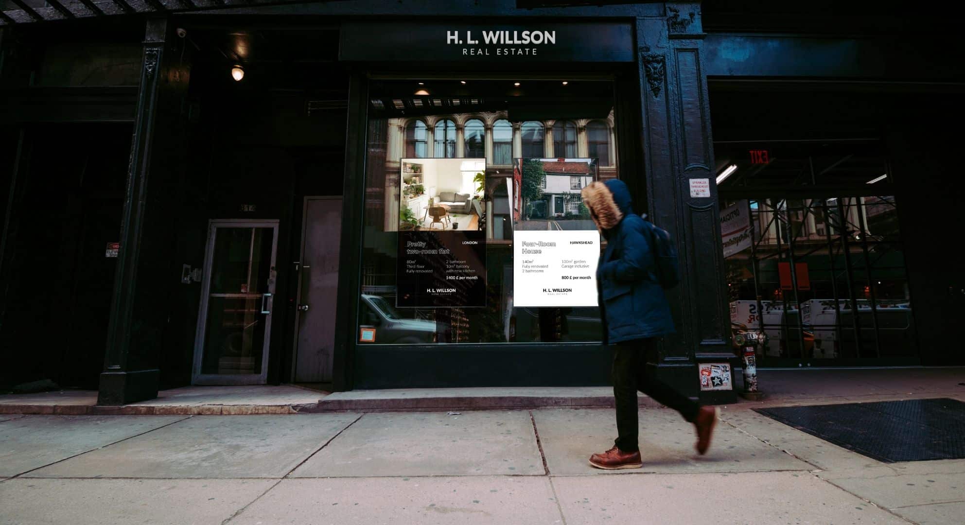 A person walks past the H. L. Willson Real Estate office, located in a building with dark exterior walls and large windows displaying listings created through small format printing.