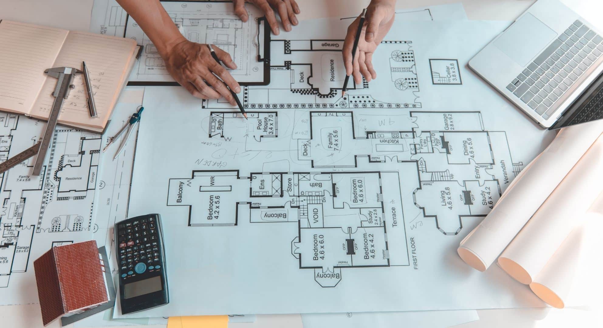 Two individuals review architectural blueprints on a table with a calculator, notebook, measuring tools, laptop, and rolled-up plans. They discuss how 3D printing could be integrated into their designs to enhance detail and accuracy in construction models.