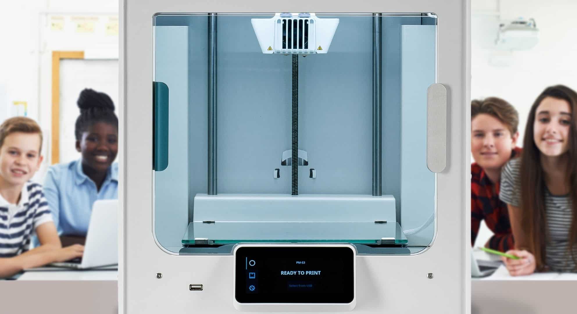 A 3D printer is in the foreground with four students sitting at a table in the background, observing intently. The printer's display screen reads 