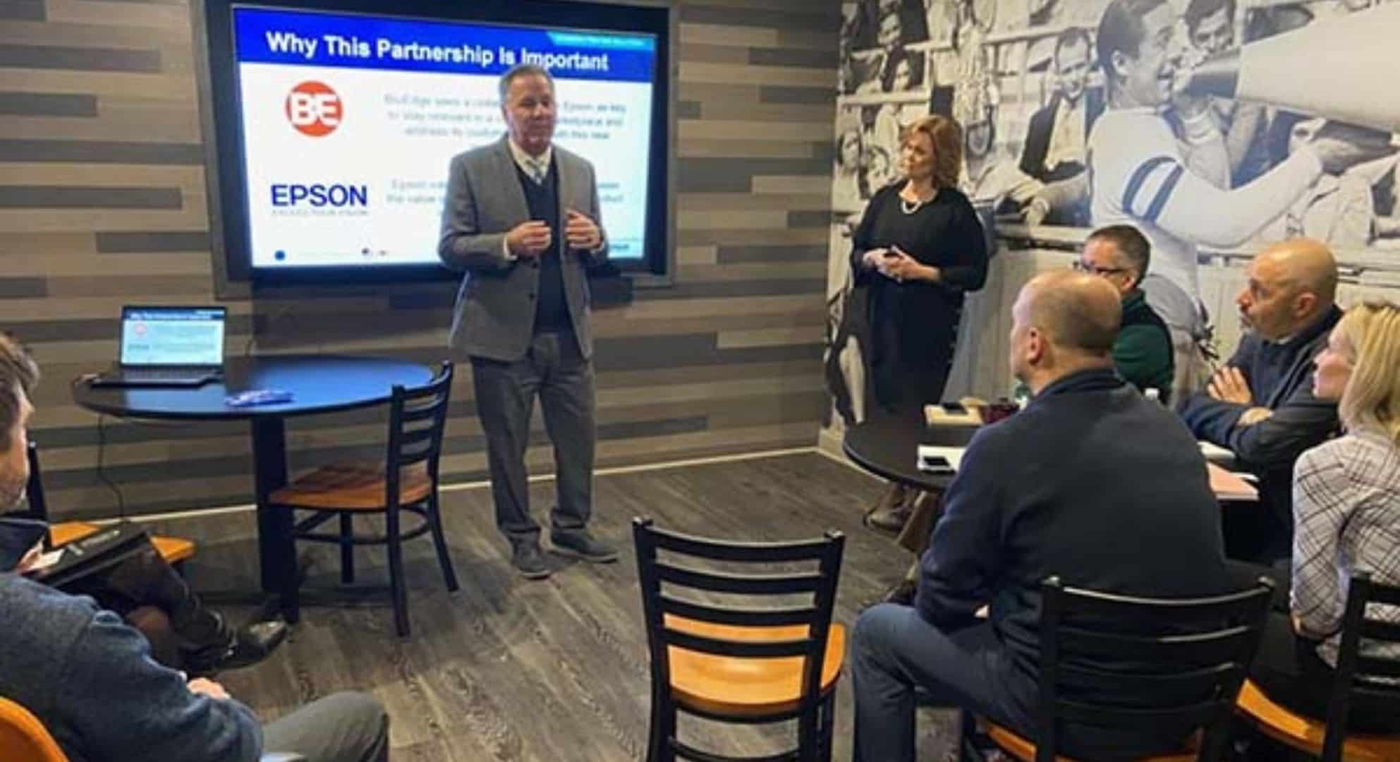 Two people stand and present to a seated audience in a room, with a screen displaying a presentation slide titled 