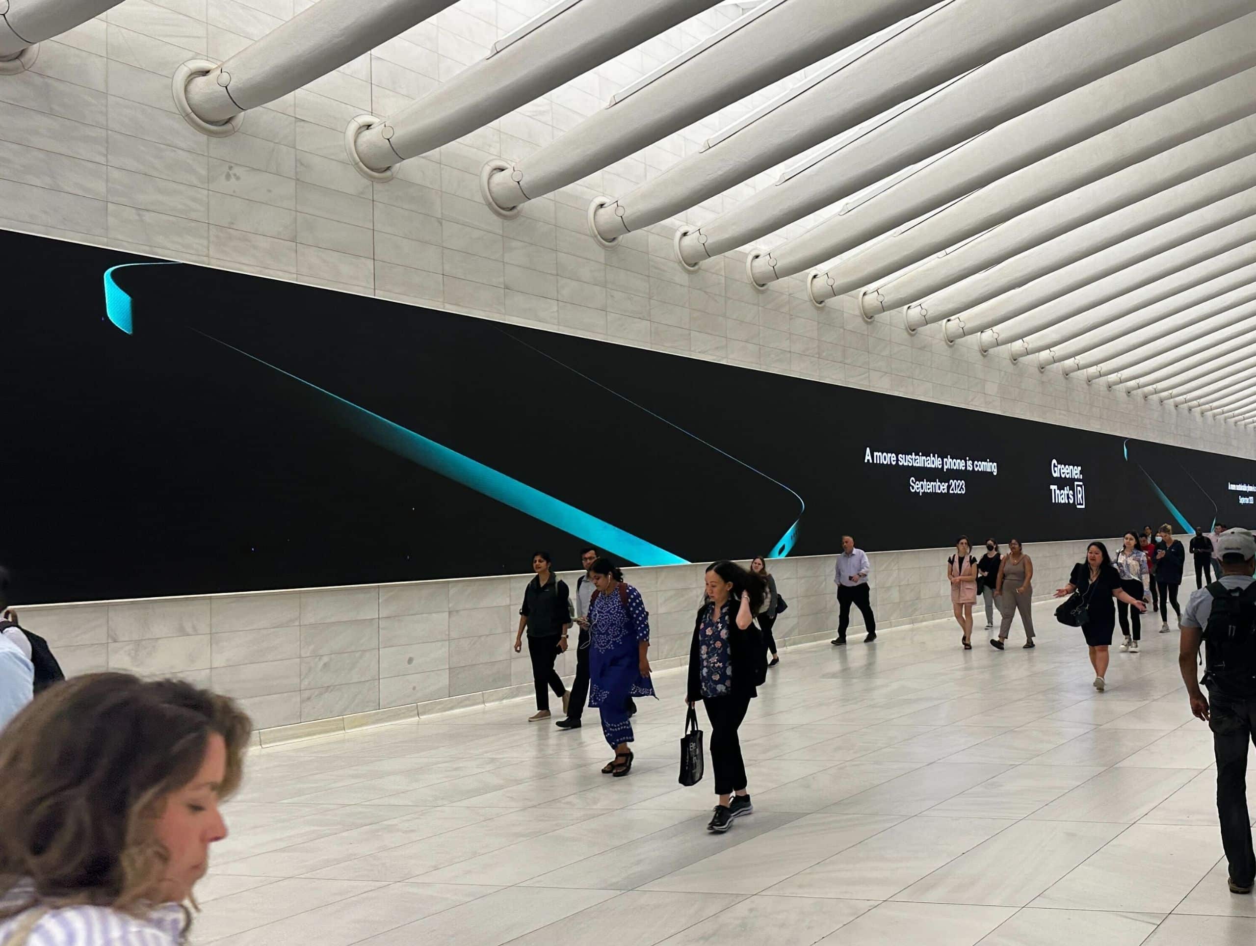 People walking in a spacious, modern hallway with a large digital billboard displaying an advertisement for a new phone launching in September 2023, made even more captivating through large format printing.