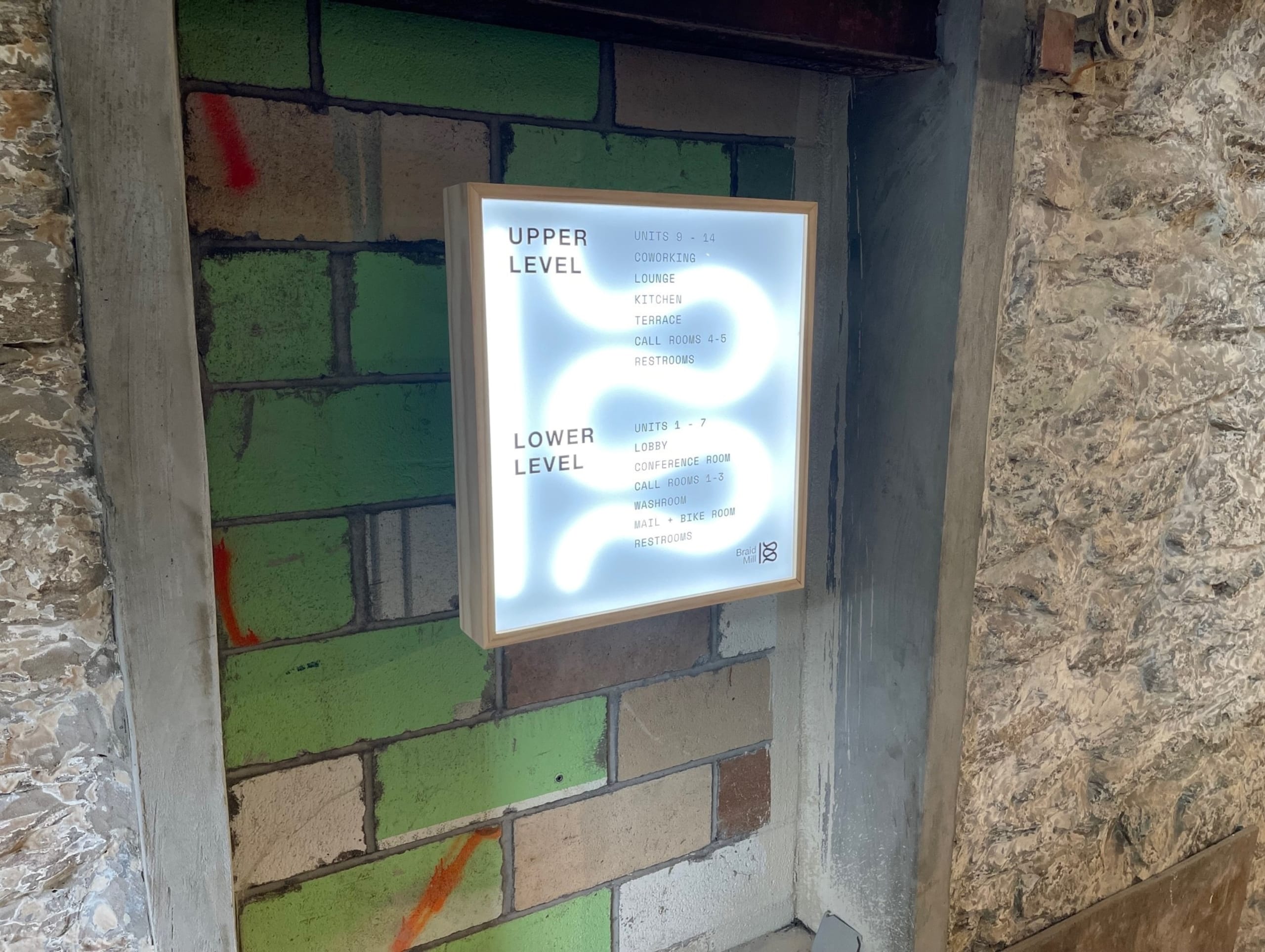 Illuminated directory sign mounted on a wall, displaying directions for different areas on the upper and lower levels of a building. The wall, featuring a combination of green and brown bricks with graffiti, includes details achieved through 3D printing.