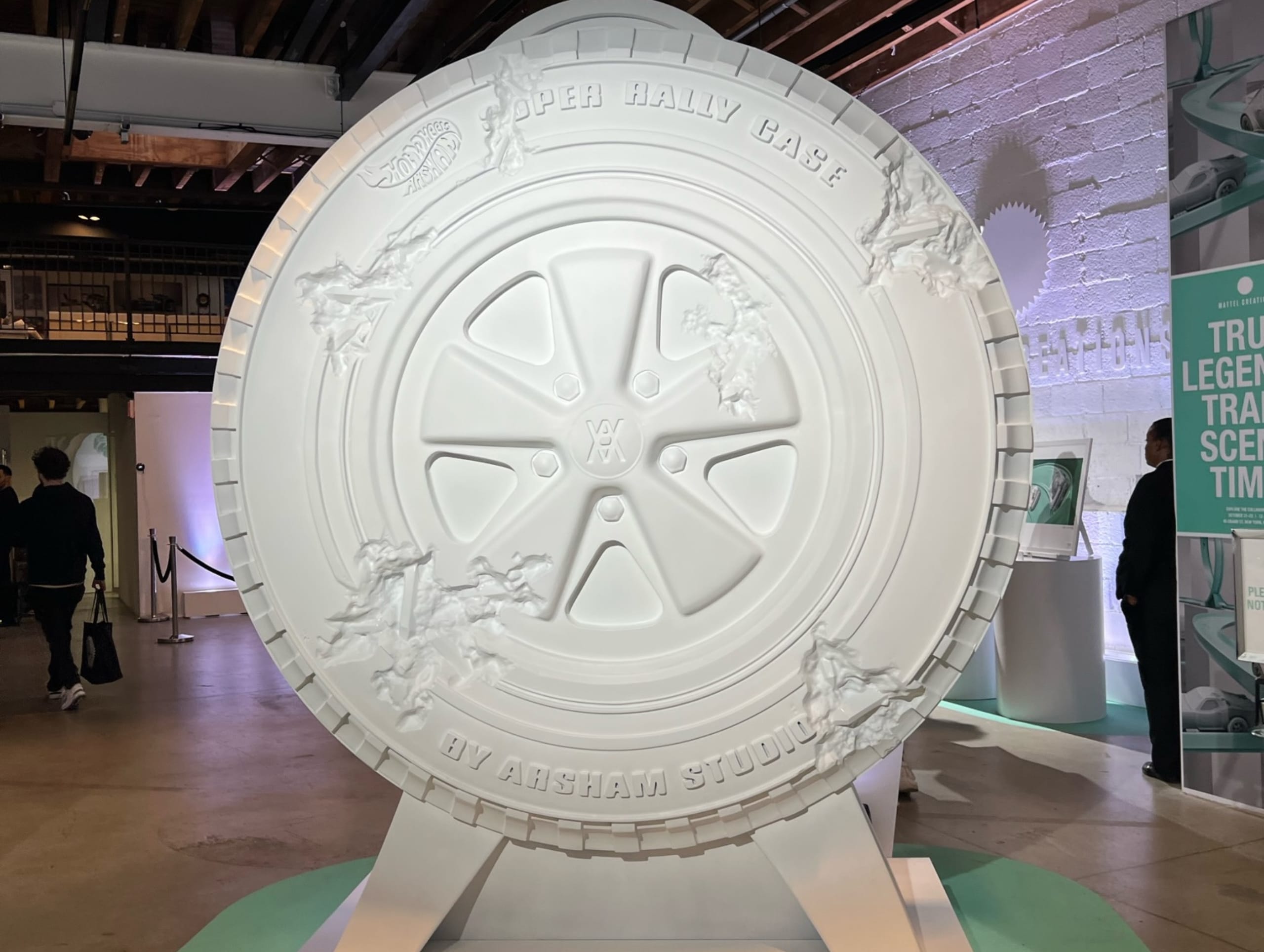 A large, white, wheel-shaped sculpture with embossed details is on display. The inscription reads "Super RALLY CASE BY ARSHAM STUDIO." Created using 3D scanning and large format printing, the exhibit is set in an industrial-style room with exposed beams.