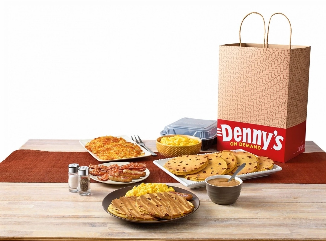 A table set with Denny's takeout, including pancakes, scrambled eggs, hash browns, sausage, bacon, and a brown paper bag marked "Denny's On Demand," sits ready for a delicious breakfast. It's like 3D scanning your perfect morning feast.