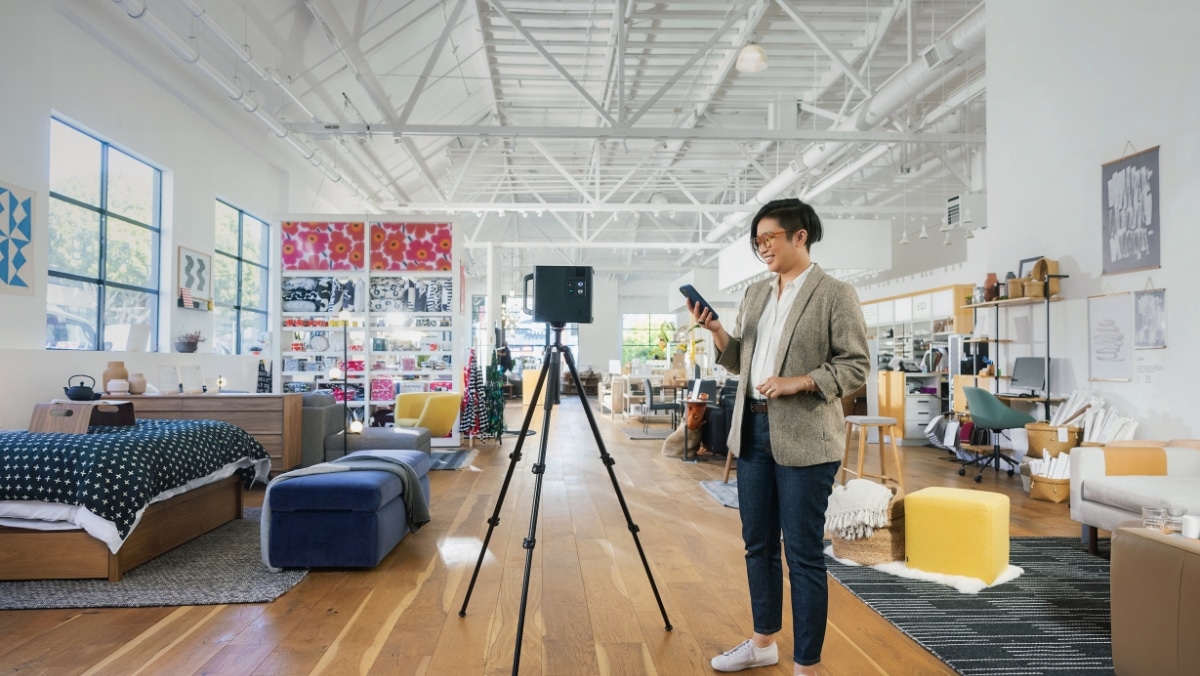 A person stands next to a tripod-mounted device in a spacious, brightly lit showroom displaying various furniture and decor items, perhaps utilizing 3D scanning technology for precise measurements.