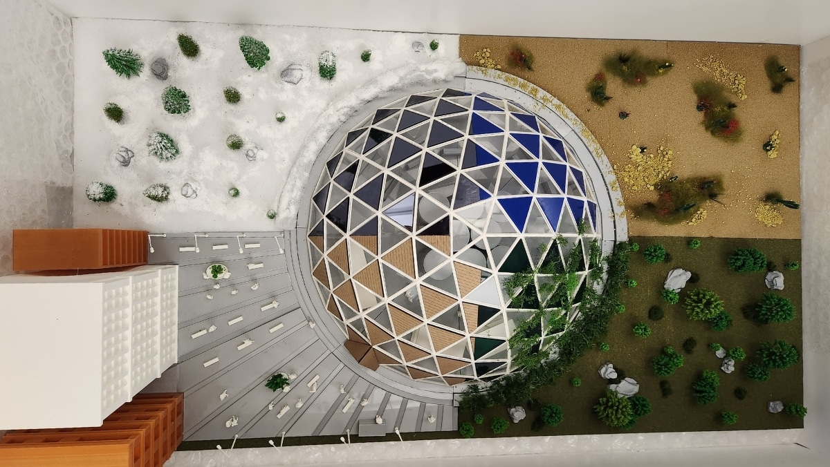 A top-down view of a geodesic dome model, created via 3D printing, is surrounded by diverse landscapes: snow and coniferous trees on the left, a grass area with shrubs on the right, and a mix of buildings and pathways nearby.