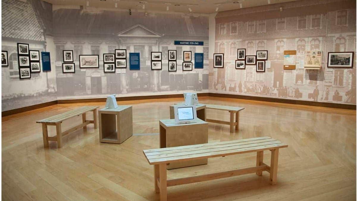 A museum exhibit room with wooden benches, two small display cases, and framed photos on the walls arranged in rows. The floor is wooden, and subdued grayscale wall art depicts historic buildings. One display features models made using 3D printing to bring historical structures to life.