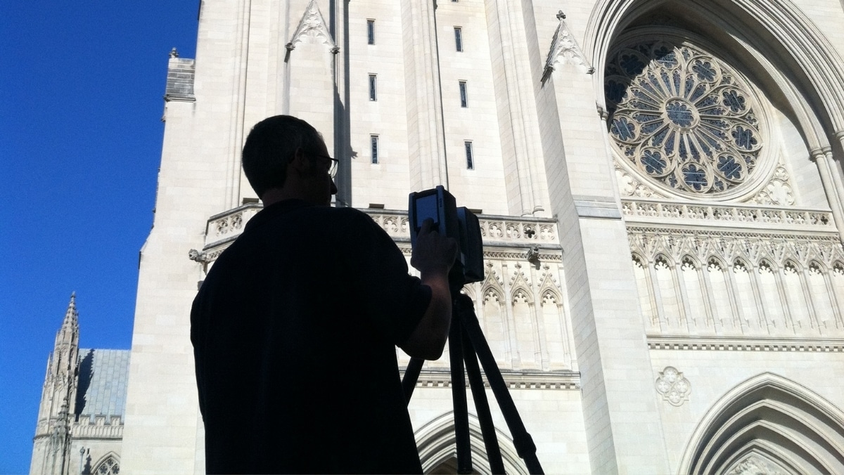 A person stands with a camera on a tripod, photographing an ornate, gothic-style cathedral facade under a clear blue sky, possibly planning to use 3d scanning technology to create a detailed virtual model.