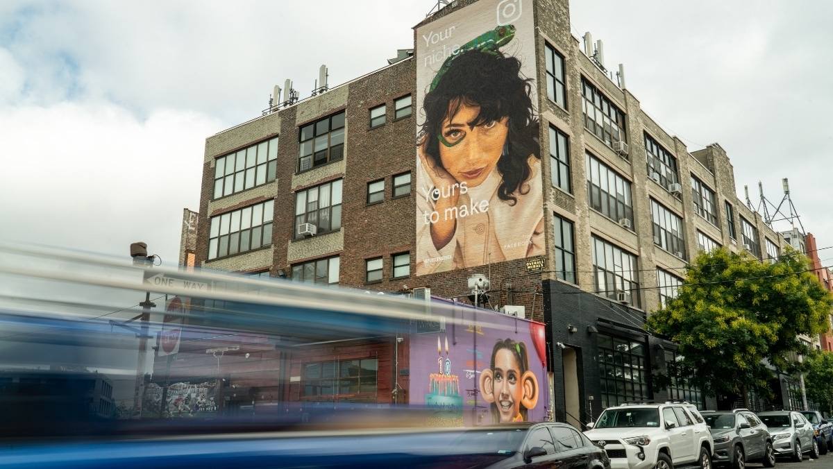 A bus passes by a multi-story brick building adorned with a large mural of a woman’s face and the text "Yours to make," capturing the innovative spirit of web to print services.