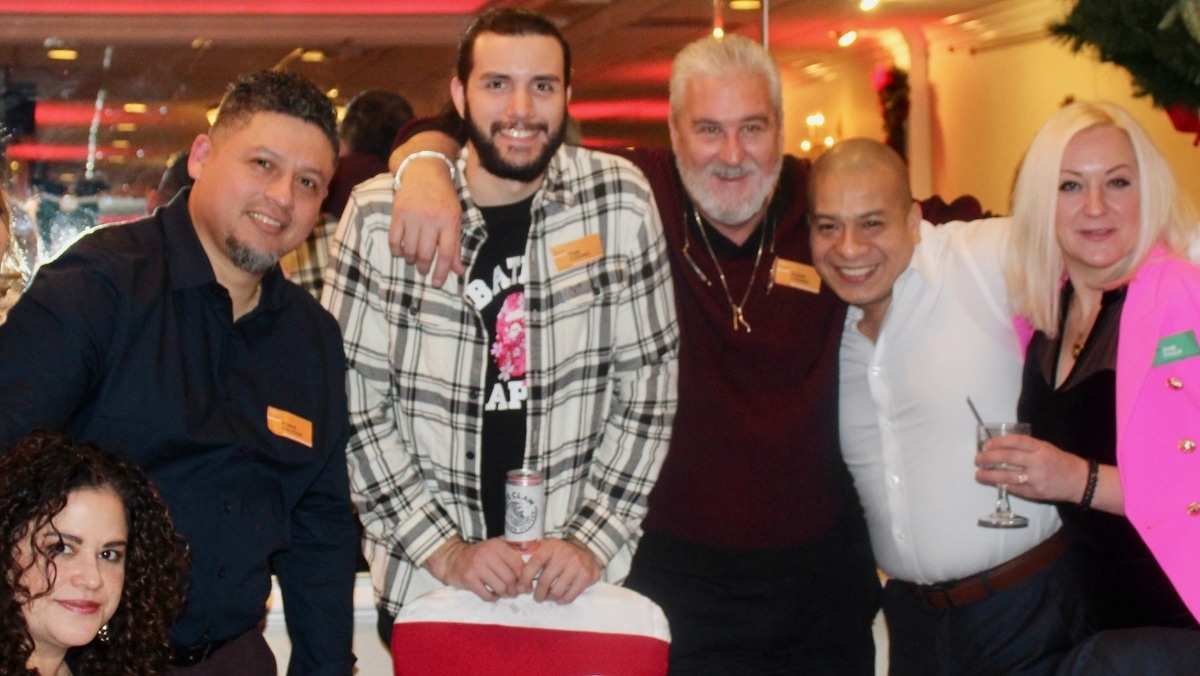 A group of five adults, some standing and some seated, smile at the camera during a social gathering, reminiscent of a snapshot captured through large format printing. One person holds a drink, another has their arm around the person next to them.