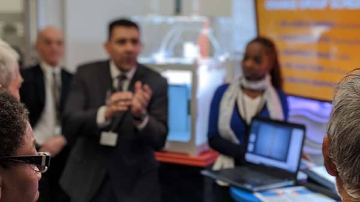 A group of people listening to a presentation. A man is speaking while others listen, and a woman stands beside a laptop showing a screen. An orange screen with text is visible in the background, highlighting advancements in 3D scanning and large format printing techniques.