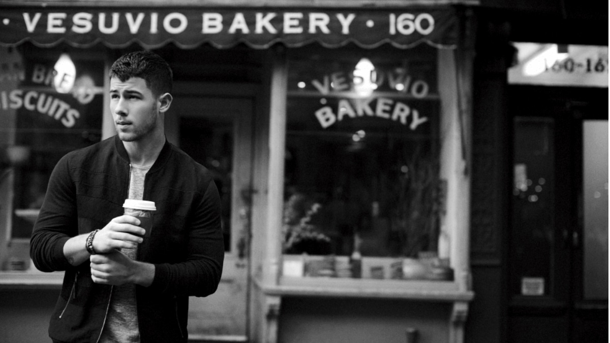 A man holds a coffee cup while standing in front of Vesuvio Bakery, its charming facade displaying a sign that reads "VESUVIO BAKERY 160." The man, dressed in casual clothing, seems to be admiring the intricate design of the sign as if it were produced through small format printing.