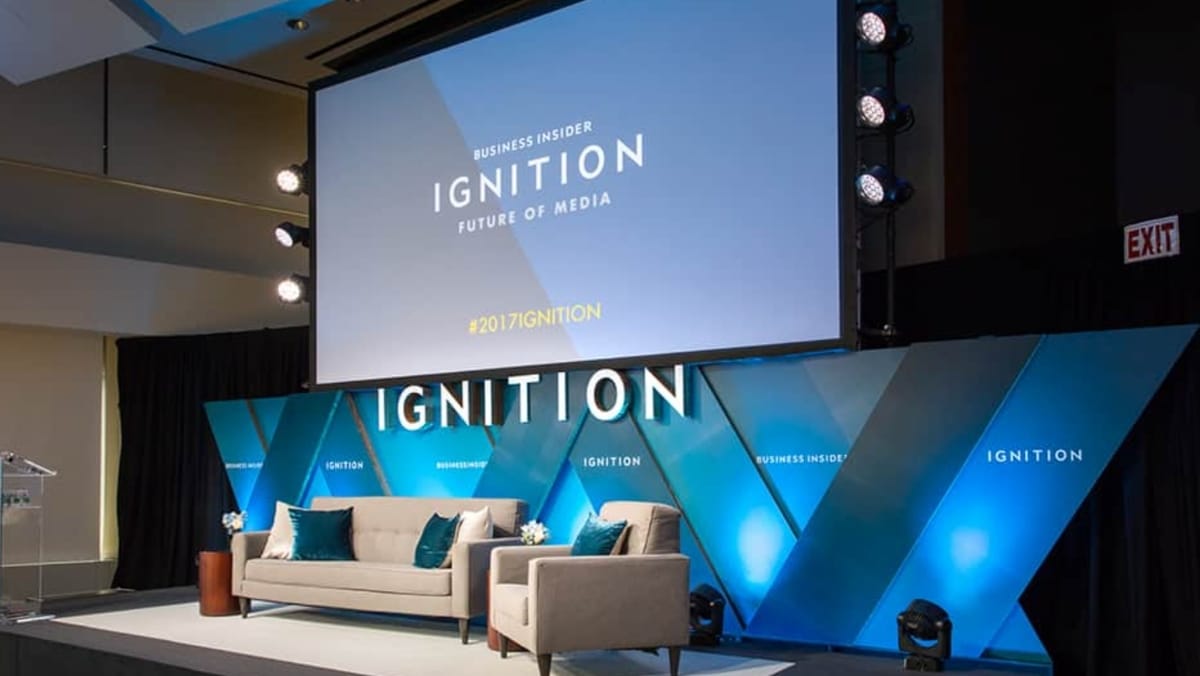 A stage setup for the Business Insider Ignition: Future of Media conference, featuring a large screen enhanced by 3D printing, blue accents, and two gray sofas in front.