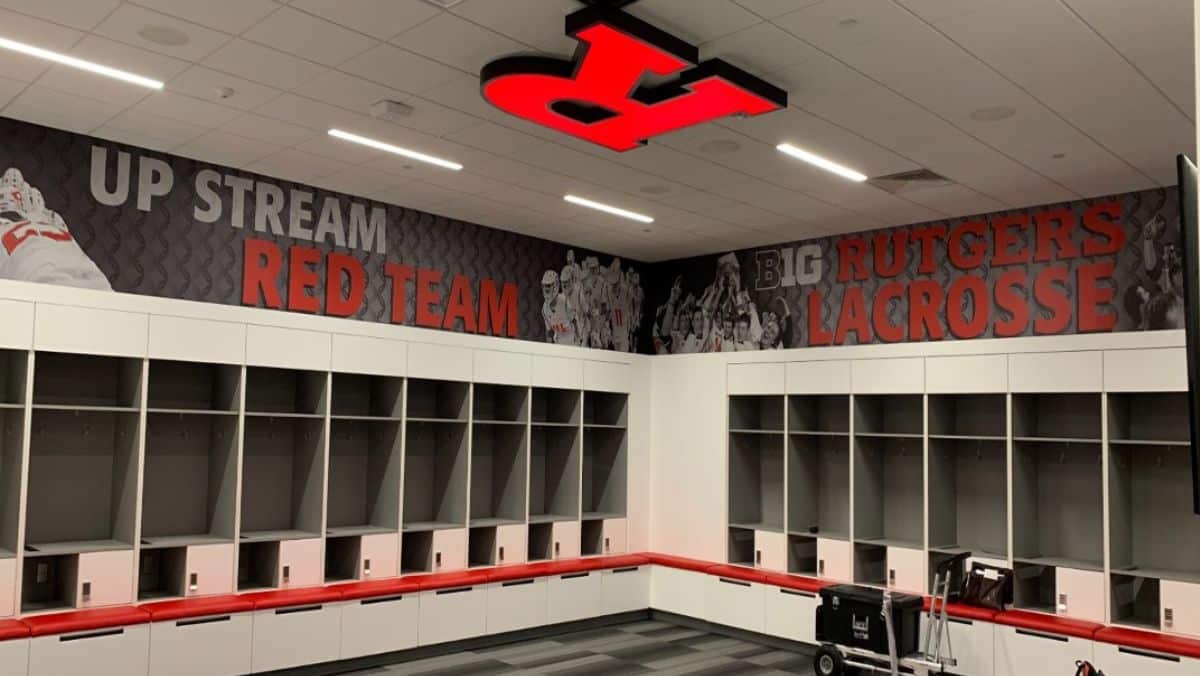 A lacrosse locker room with red cushioned benches, empty cubbies, and a large red 'R' on the ceiling. The walls display motivational text for the Rutgers lacrosse team, enhanced by large format printing. An equipment cart rests in the foreground.