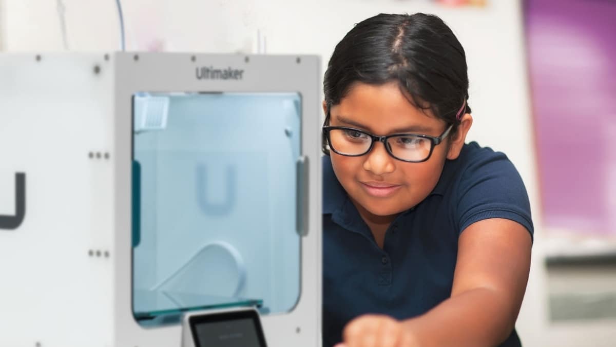 A student wearing glasses and a navy blue shirt operates a 3D printer in a classroom, showcasing the intricate possibilities of 3D printing technology.