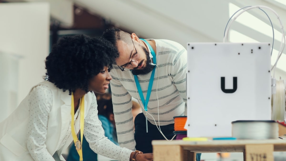 Two people are collaborating and observing a 3D printer in action, with colorful filament spools visible on a table next to the printer, showcasing the impressive capabilities of 3D printing.