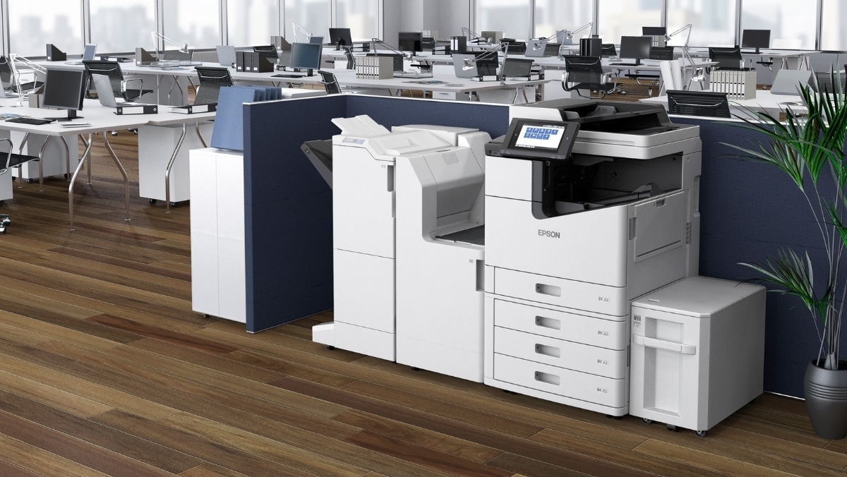 A modern office with empty cubicles and desks features a large Epson printer specialized in small format printing against a partition wall on a wooden floor, along with a potted plant nearby.