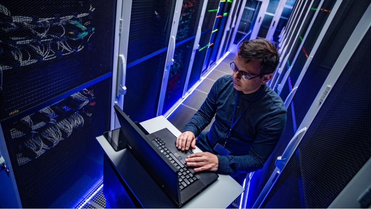 A person working at a laptop in a data center, surrounded by server racks with illuminated lights and large format printing equipment. The space is dimly lit with blue accent lighting.