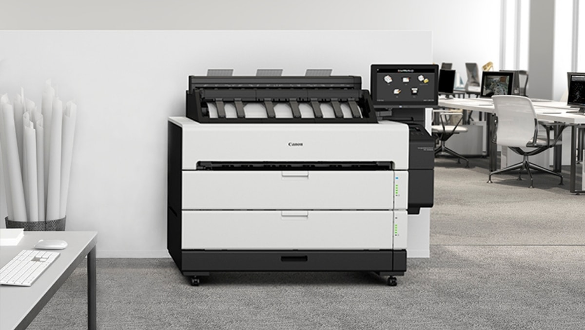 A large multifunction printer, capable of small format printing and 3D scanning, sits in an office space with desks and chairs in the background. Several rolled-up papers are visible on a nearby shelf.