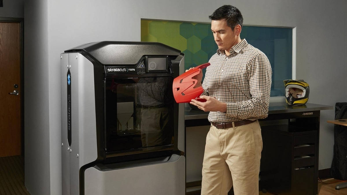 A man inspects a red object he has just taken out from a Stratasys F123 Series 3D printer in an office setting, marveling at the precision that complements their small format printing projects.