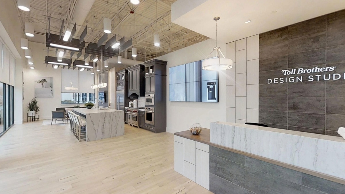 Modern design studio with an open kitchen and dining area, featuring sleek, contemporary decor, large marble island, and wood flooring. A reception desk is in the foreground with "Toll Brothers Design Studio" signage. The space also showcases cutting-edge 3D printing displays for innovative projects.