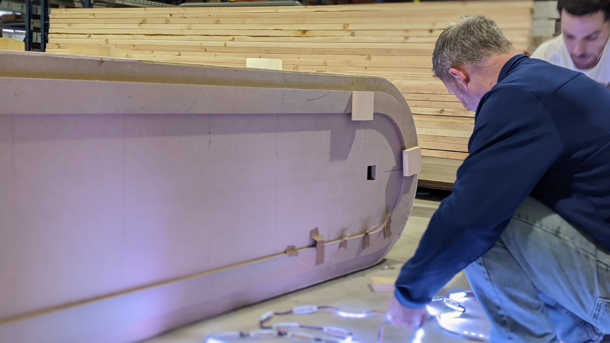 Two people are installing LED strip lights on a large, curved wooden structure in what appears to be a workshop, possibly preparing it for a web to print display.