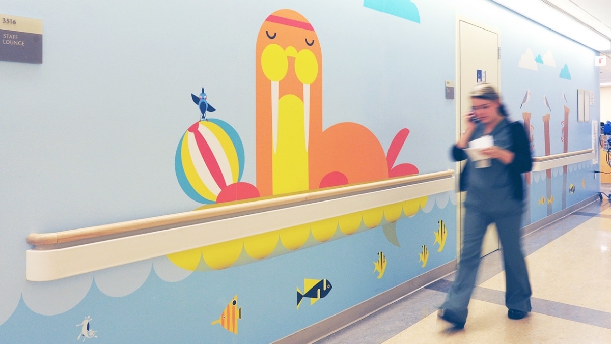 A hospital staff member walks past a colorful mural featuring a walrus, a beach ball, and various sea creatures on a light blue wall, showcasing the intricate detail achievable through large format printing.