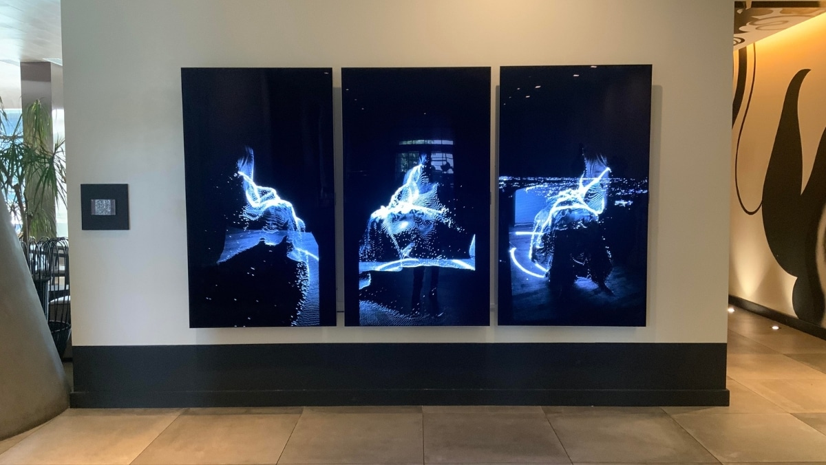 Three vertical digital screens are mounted on a wall, displaying abstract light patterns. The screens are illuminated in a dark setting, enhancing the vibrant white and blue light streaks, reminiscent of detailed textures one might achieve with 3D printing.