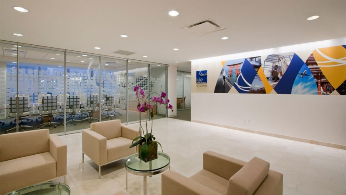 Modern office lobby with beige seating, glass walls, and a colorful mural. A small round table holds an orchid plant. Conference room visible through glass partition, where a 3D printing display showcases innovative designs.