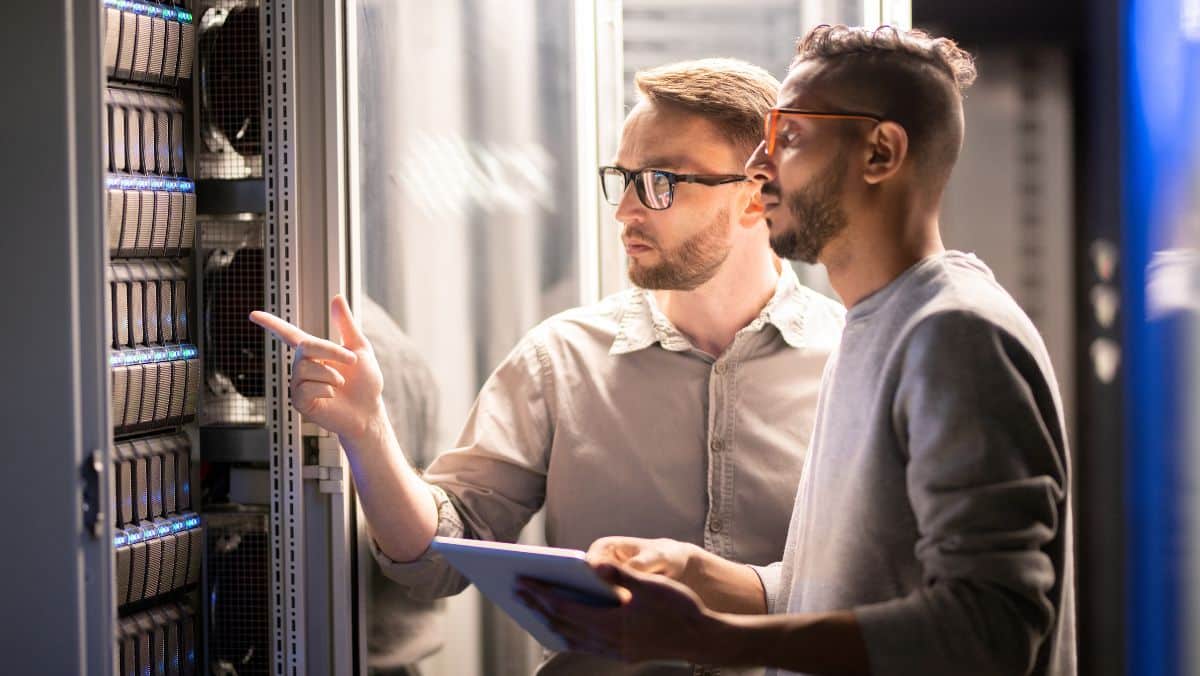 Two men stand in a server room, one pointing at a server rack while the other holds a tablet. They appear to be discussing the equipment and its integration with web-to-print solutions.