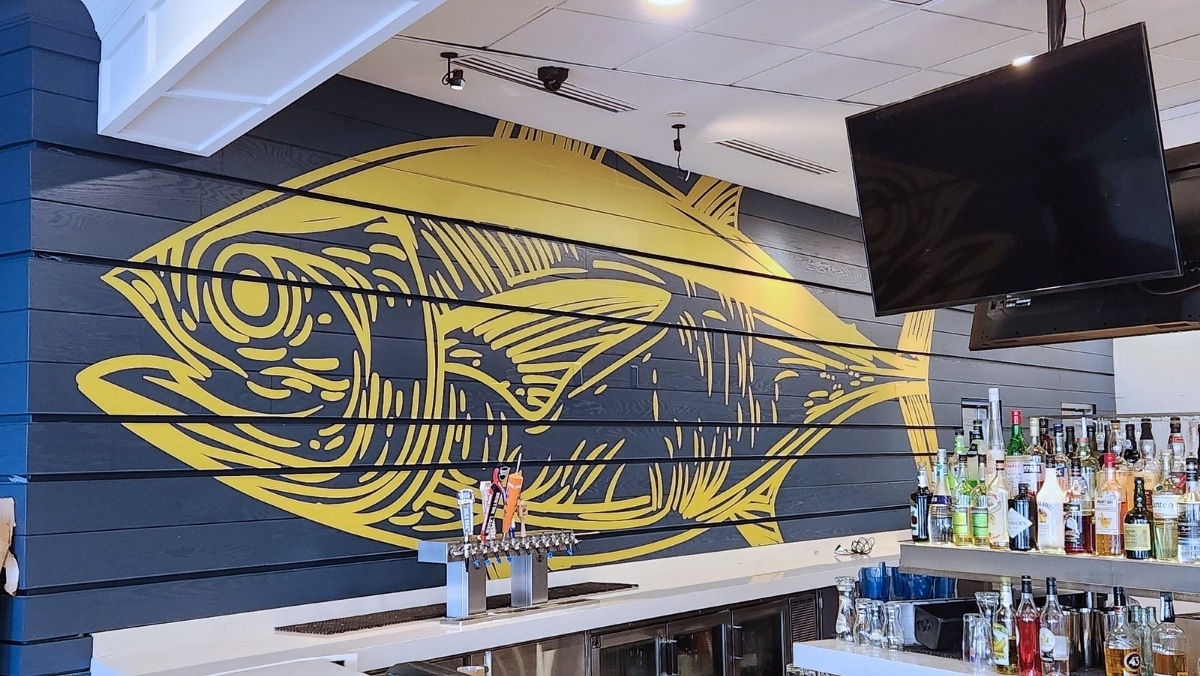 A bar interior with a yellow fish mural on a navy blue wooden wall, created using large format printing, a row of taps on the counter, and various bottles arranged neatly on a shelf. A TV is mounted above the bar area.