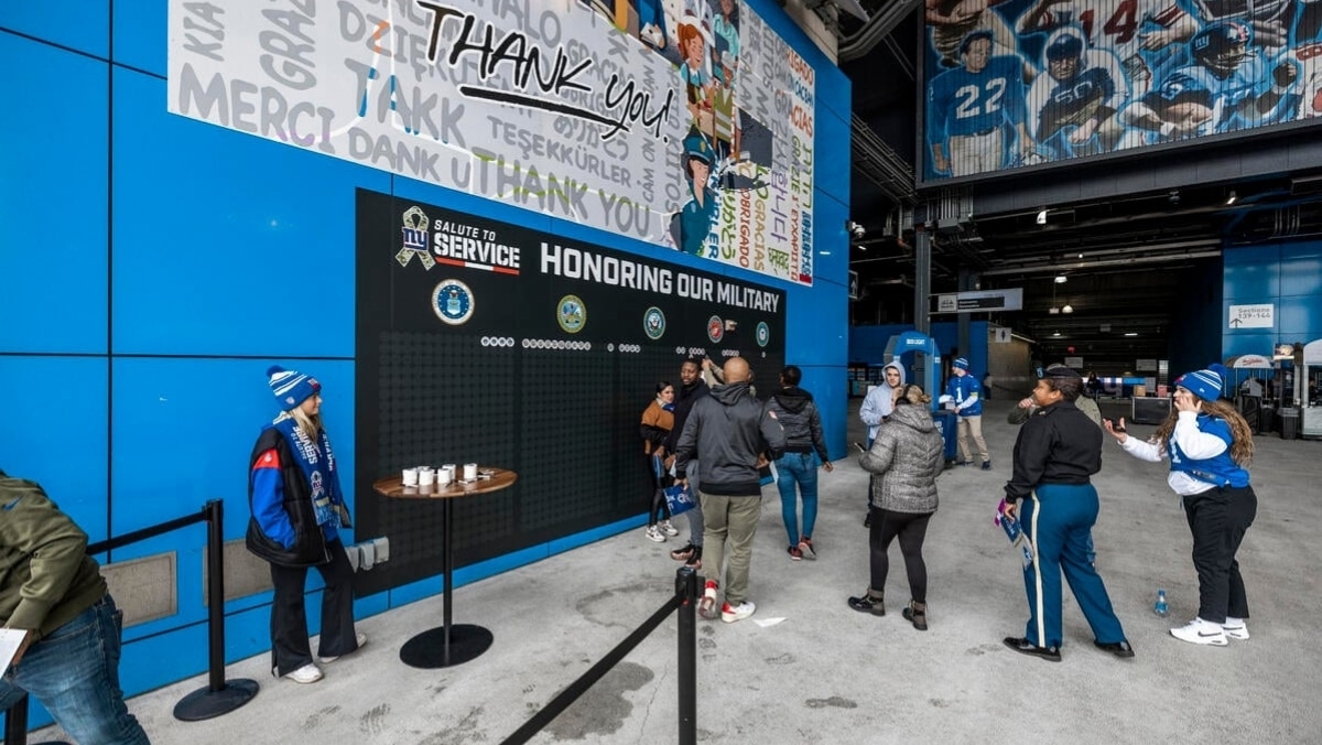 People in an indoor stadium area walk and interact near a wall with military logos and a "Thank You" sign. The area boasts decorations, a banner honoring military service, and displays produced through large format printing.