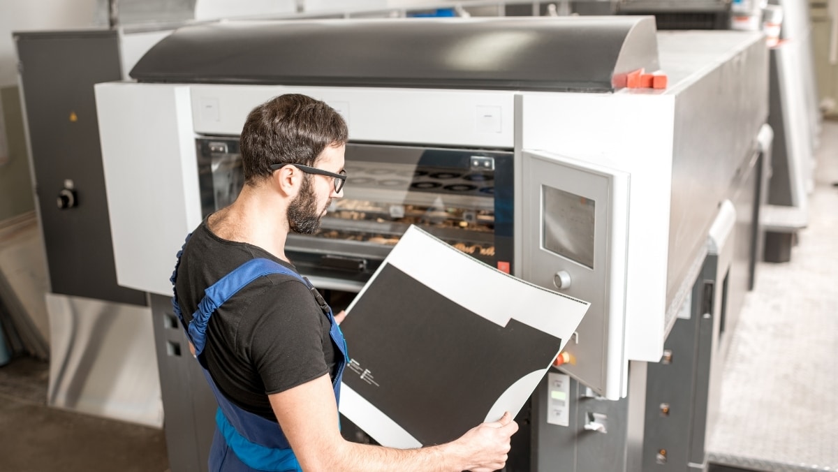 A man in blue overalls inspects a large sheet of paper in front of an industrial large format printing machine.