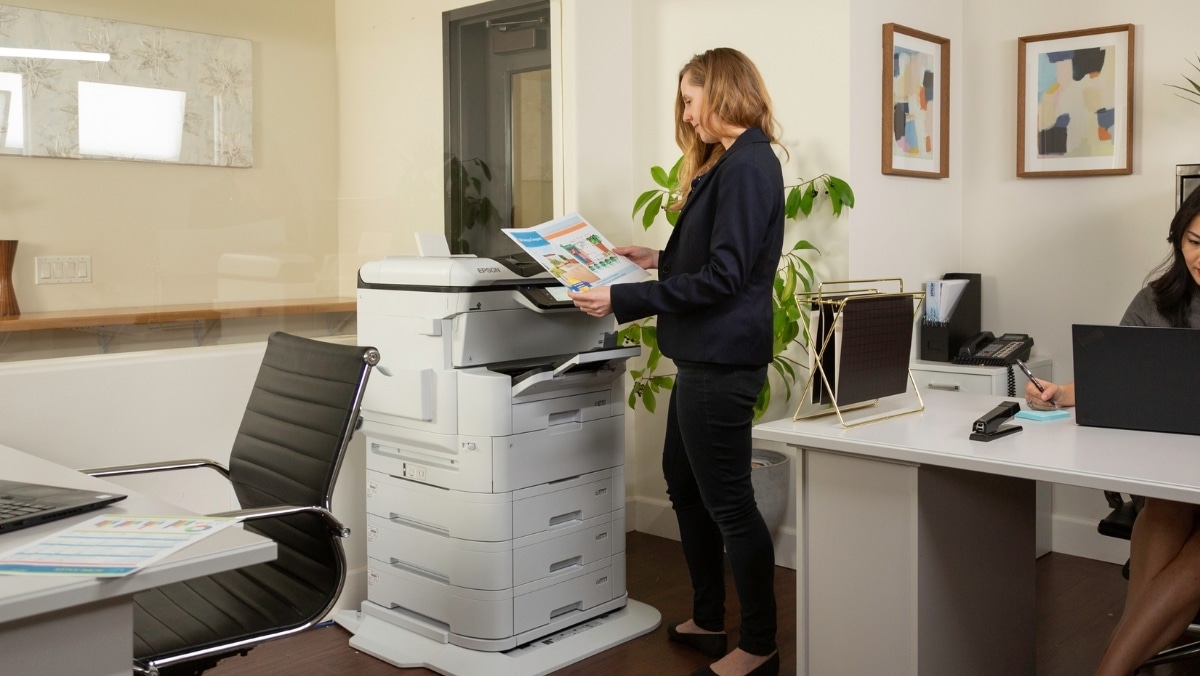 A woman stands at a photocopier printing documents, while another woman works at a desk on a laptop and phone in an office setting, where 3D scanning technology is also in use.