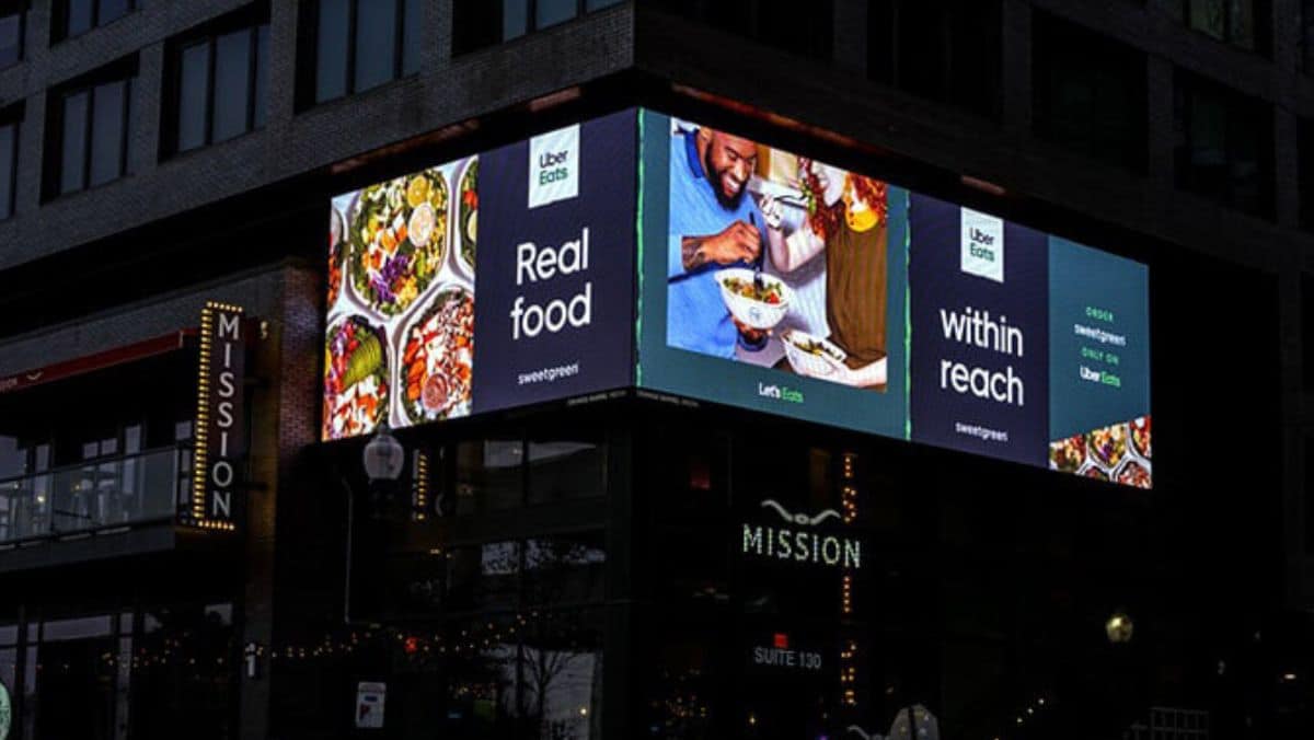 An outdoor billboard for Uber Eats and sweetgreen in a city displays images of food, with the text "Real food within reach." The large format printing on the billboard is situated on a building named "MISSION.