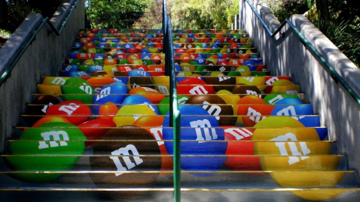 A staircase painted with large, colorful M&M candy designs on each step, giving the illusion of scattered M&Ms on the stairs, reminiscent of what you might achieve with large format printing.