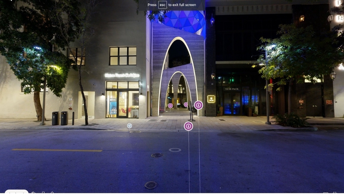 A nighttime street scene showing illuminated modern architecture with arches and trees. The building on the left has a shop with bright windows. There is a virtual tour interface with multiple points of interest, some highlighting innovative uses of 3D printing in design elements.