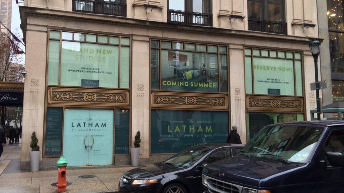 Street view of a building with multiple storefront windows advertising new luxury studios at "The Latham." Signs in the windows, created with large format printing, read "Coming Summer" and "Reserve Now," with contact information provided.