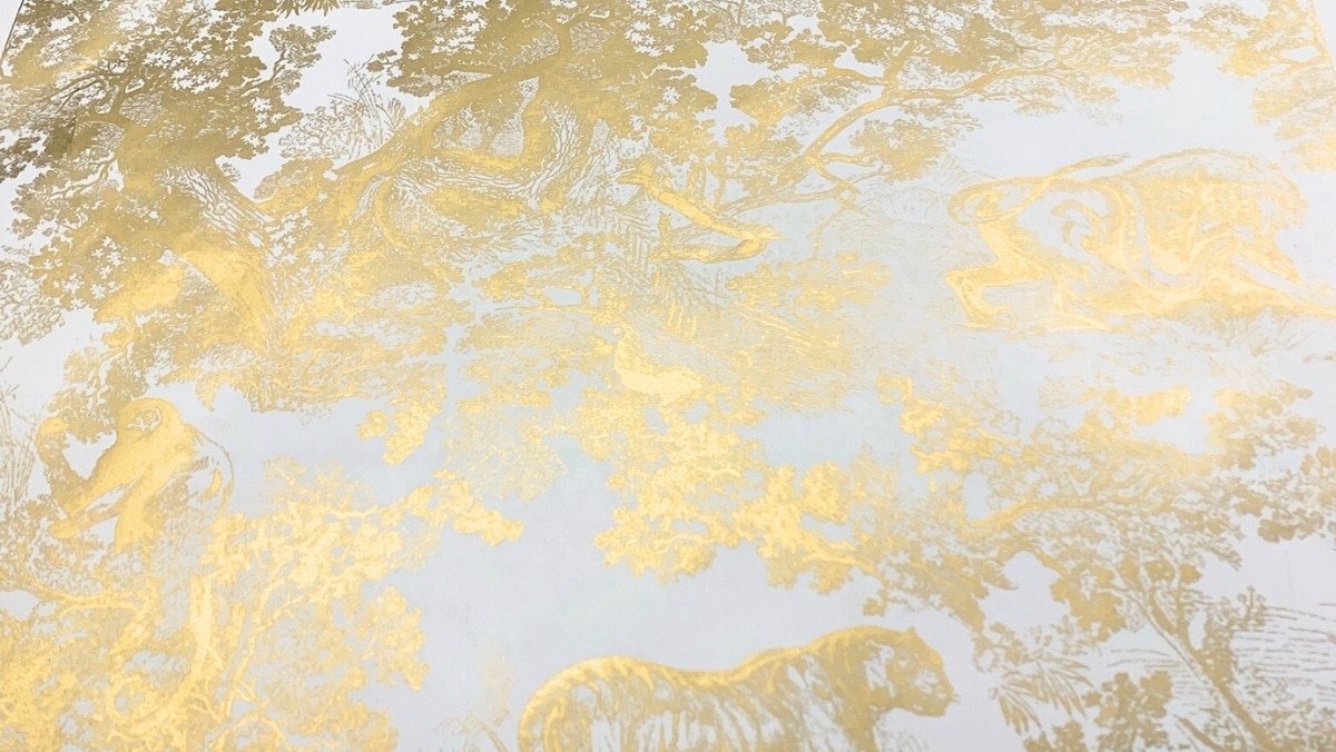 An image of wallpaper featuring intricate gold illustrations of animals, including a monkey and a lion, amidst detailed foliage and woodland scenes on a pale background, created using large format printing techniques.