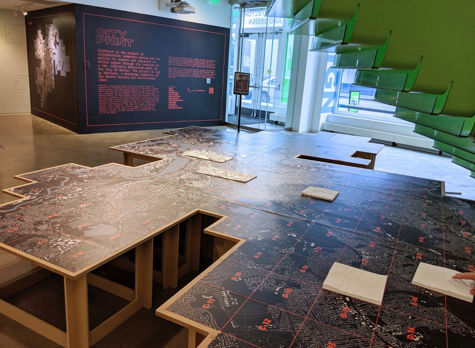 Exhibit room with a large black table featuring white and red printed maps, showcasing the possibilities of small format printing. The wall displays explanatory text, and a green staircase is visible on the right.