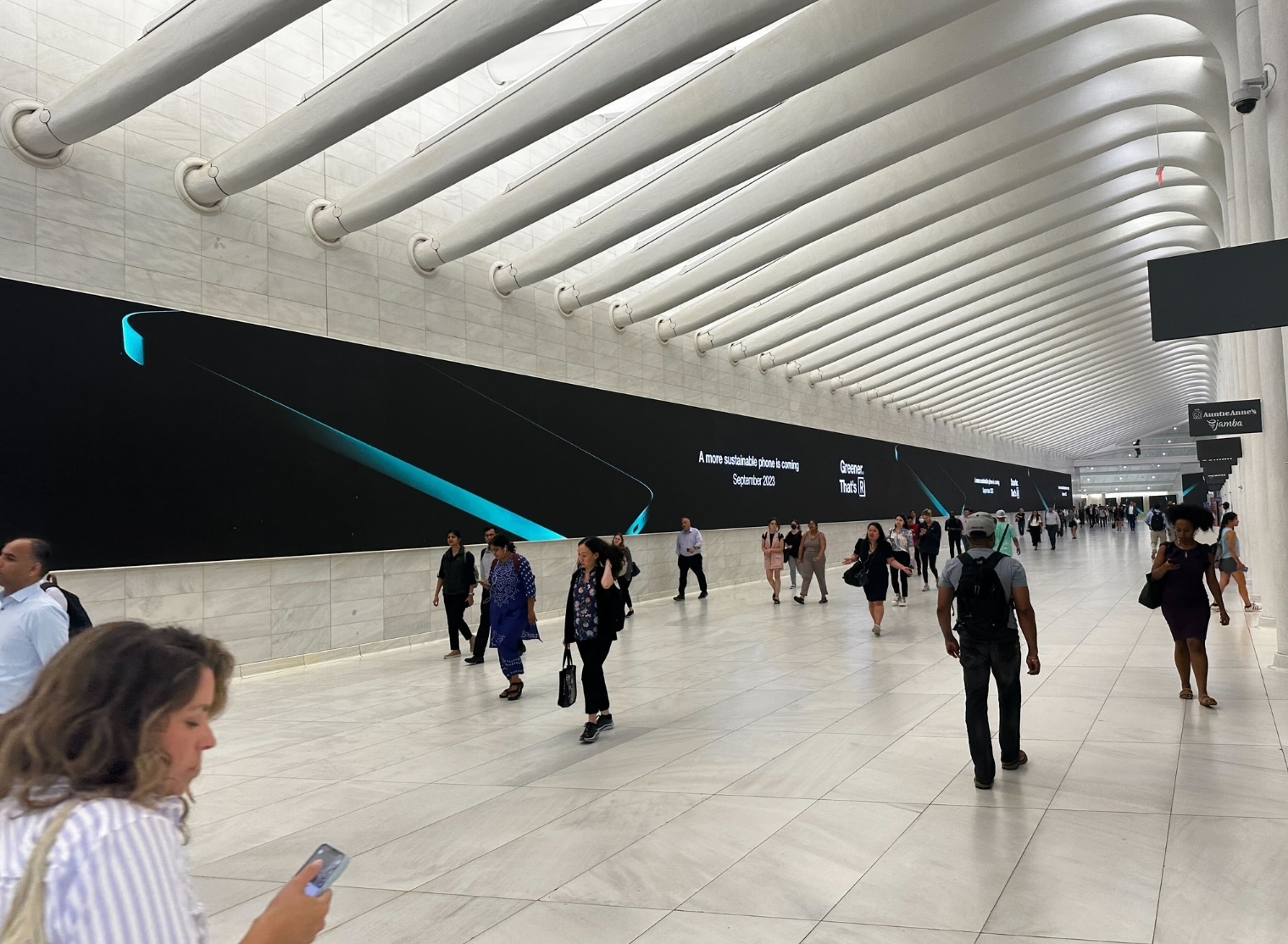 People walk through a spacious, modern corridor with a high, curved ceiling and a long black wall displaying advertisements and information. The space showcases innovative 3D printing applications that captivate the audience's attention.