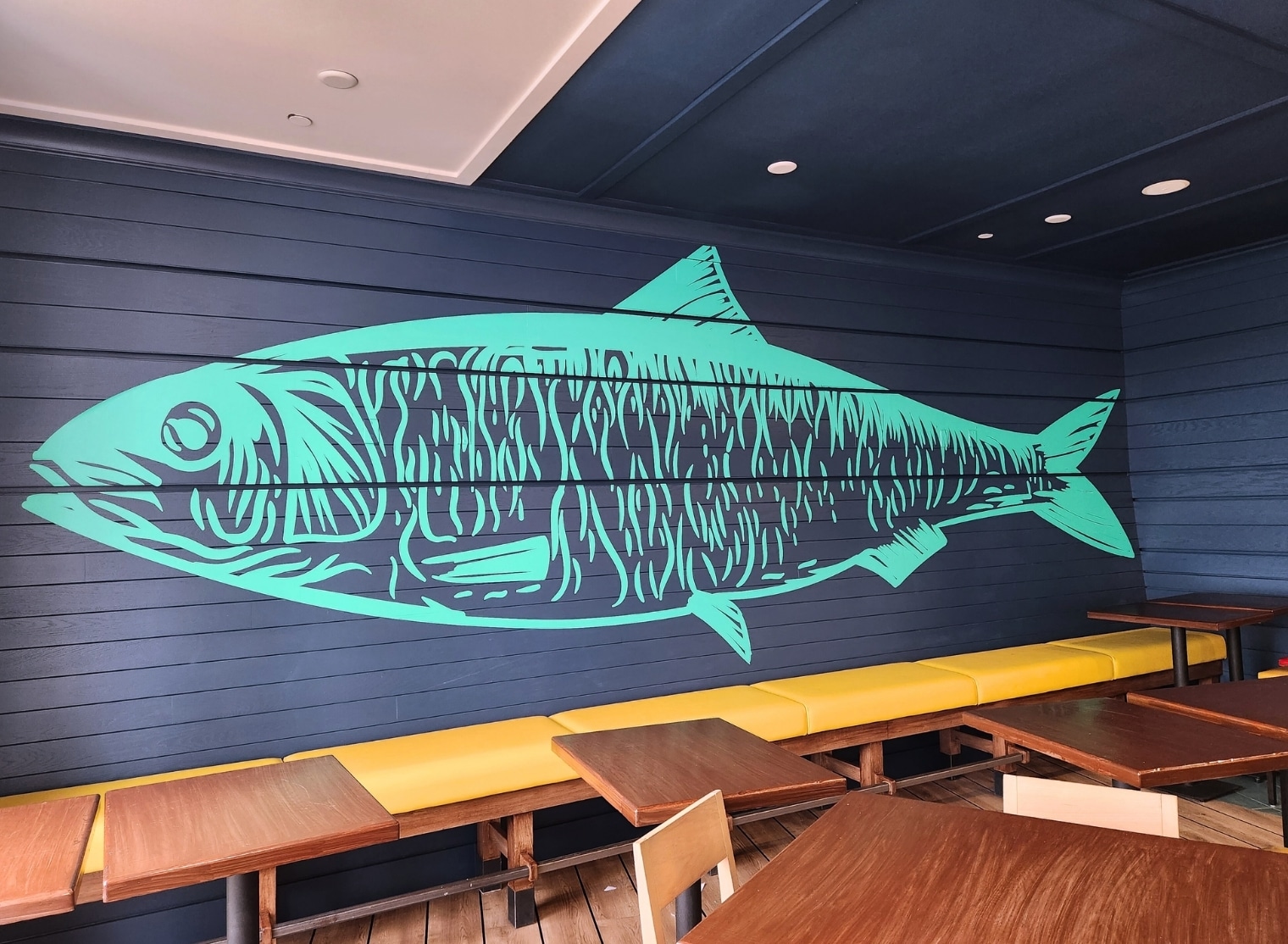 A large, turquoise fish mural is painted on a dark blue wooden wall above a yellow bench, creating an artistic seating area with wooden tables and chairs. It feels as if the vibrant scene was brought to life through the magic of 3D printing.