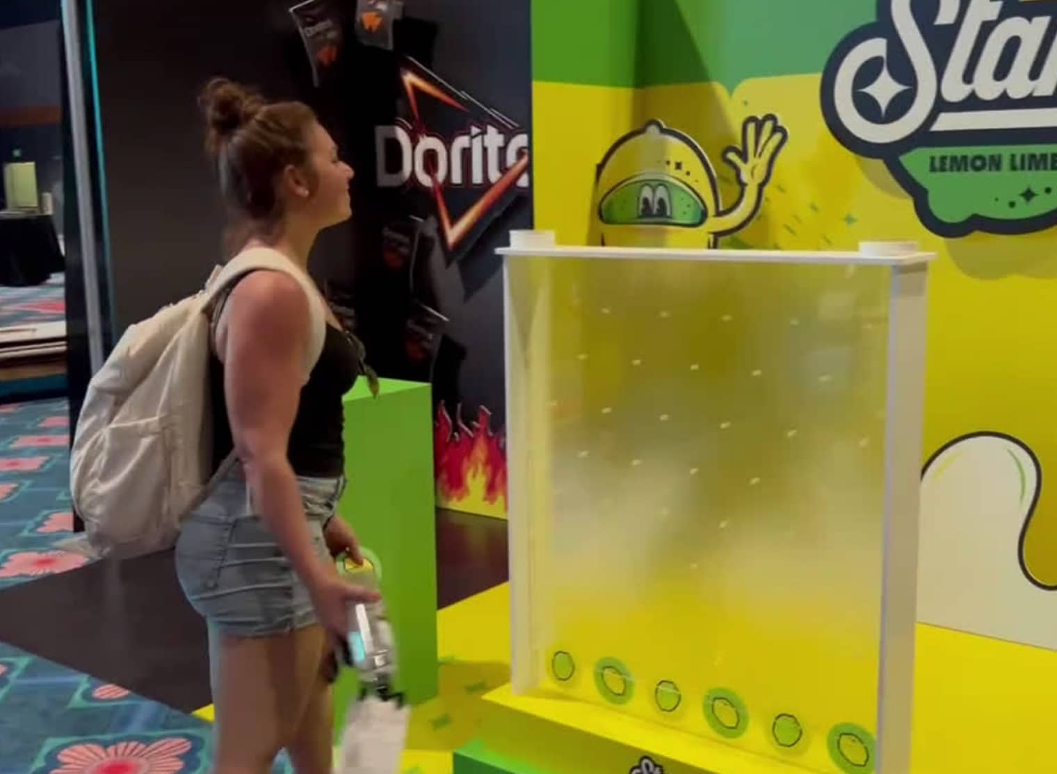 A person with a backpack and a water bottle in hand stands in front of a lemon-lime soda promotional display, enhanced by large format printing, next to a corner with Doritos branding.