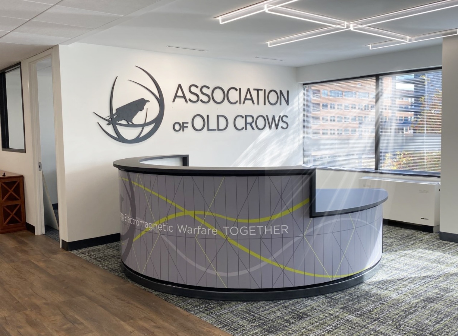 Reception area of an office with a sign reading "Association of Old Crows" on the wall and a curved desk with text promoting electromagnetic warfare. Large windows are in the background, complementing the sleek design, while large format printing ensures all signage is both clear and impressive.