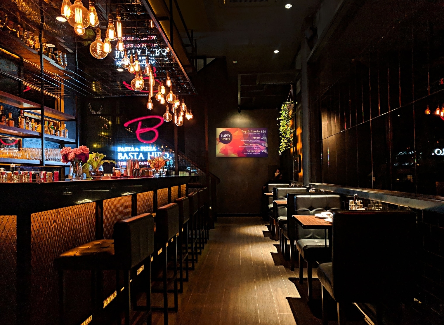 A dimly lit modern bar with hanging lights, high stools along the counter, and tables with seating on the right side. A neon sign and flower arrangement are visible near the bar area, adding a touch of charm reminiscent of small format printing details.